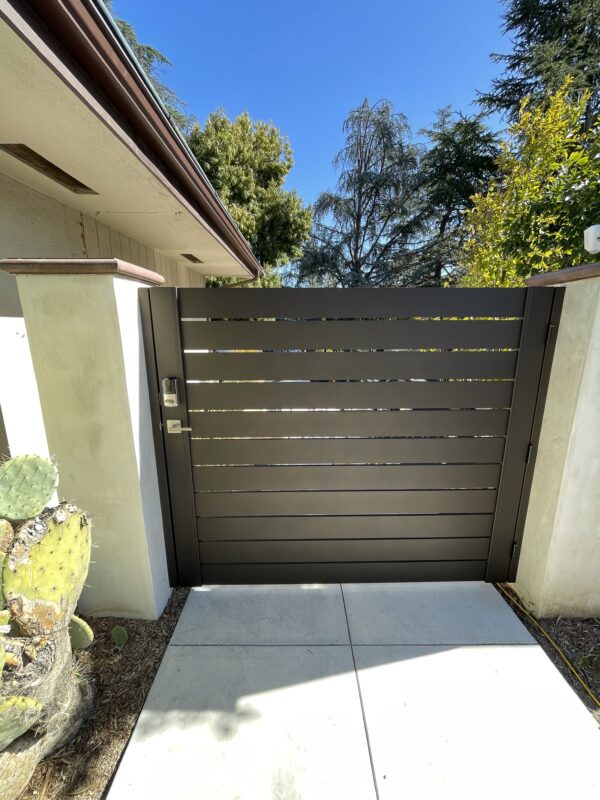 Modern bronze aluminum side yard gate installed, surrounded by greenery and concrete pathway.
