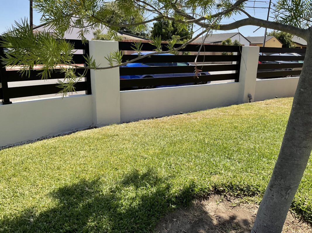 Modern black aluminum fence against a white wall with green grass and tree shadows.