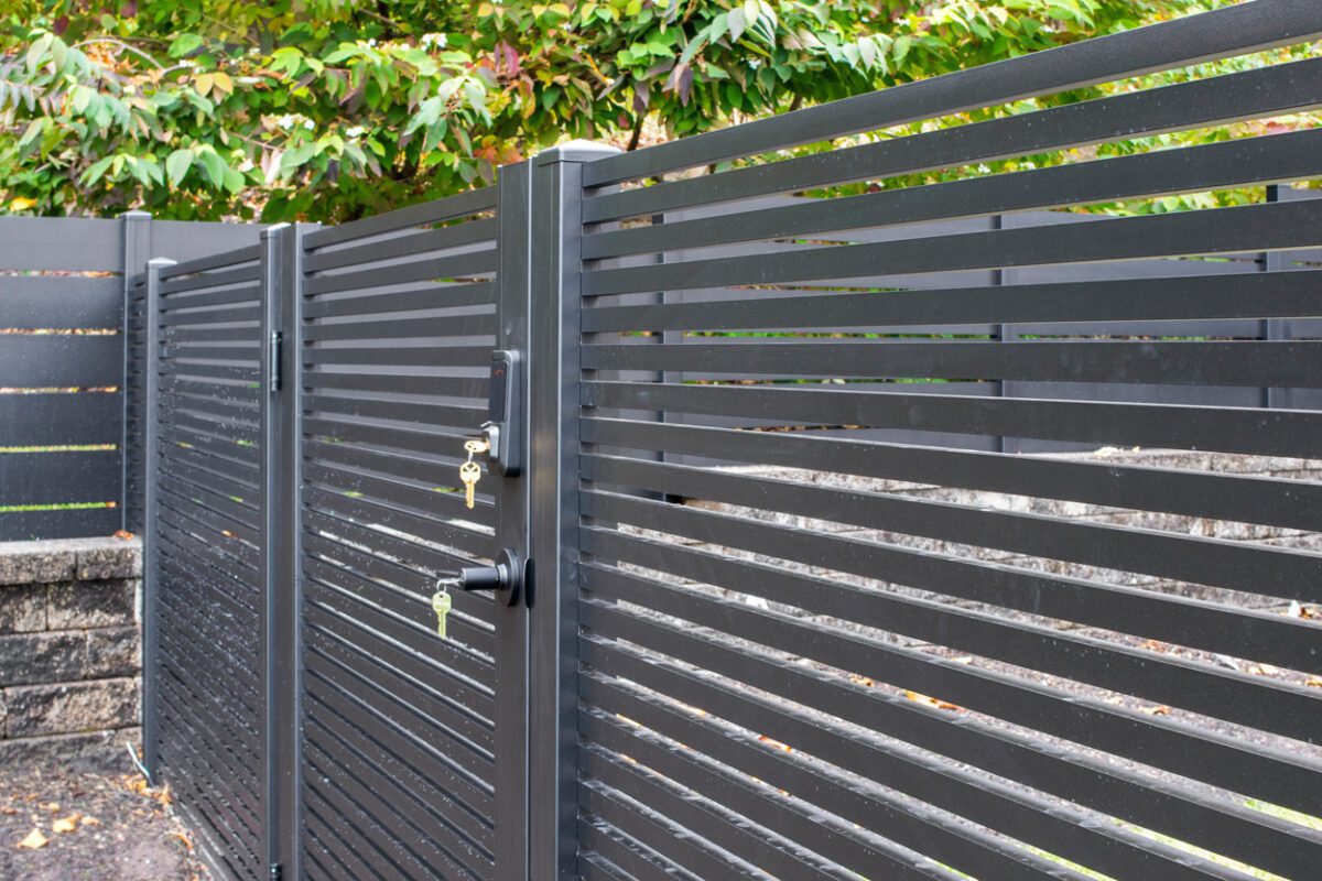 A modern black aluminum fence with horizontal slats and a lock near greenery.