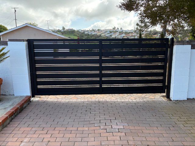 Modern black driveway gate with horizontal slats, framed by white walls and brick driveway.