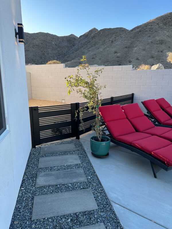 Matte black aluminum gate with slats near red lounge chairs in a desert setting.