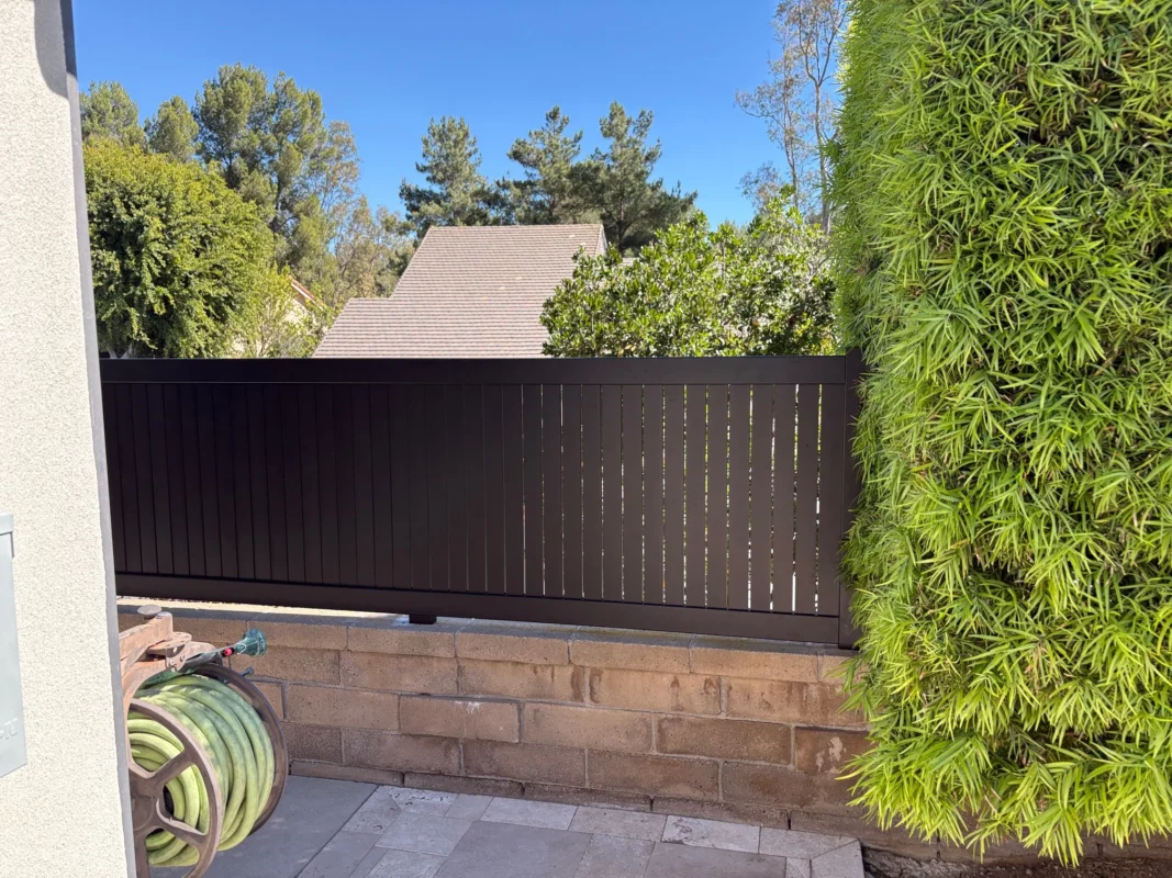 Close-up of black aluminum vertical wall topper with 2.5-inch slats and 1/4-inch gaps, installed on a block wall next to house and garden hose.