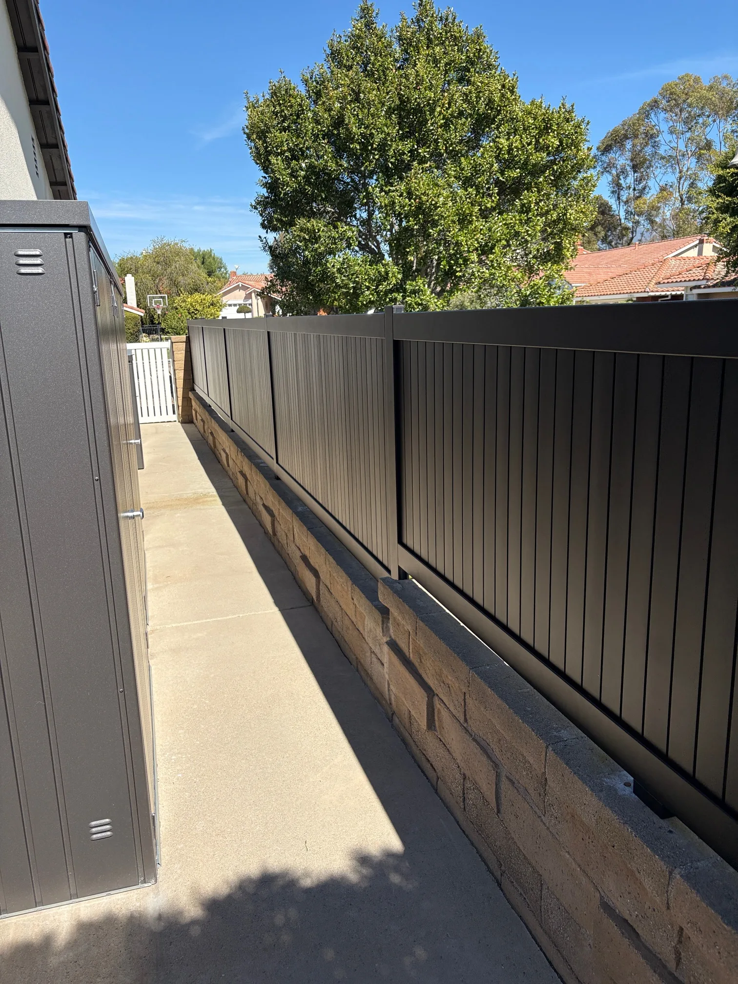 Black aluminum vertical wall topper on block wall next to walkway with trees and residential homes in background.