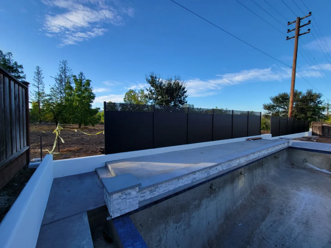 Black aluminum fence on a raised concrete base beside a pool under construction.