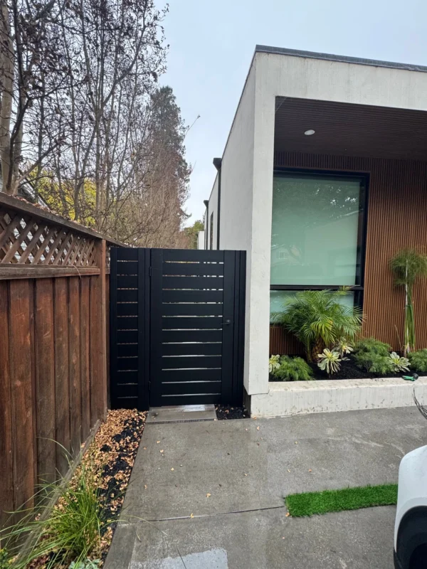 Black pedestrian gate with horizontal slats, between concrete driveway and wooden fence next to modern home facade.