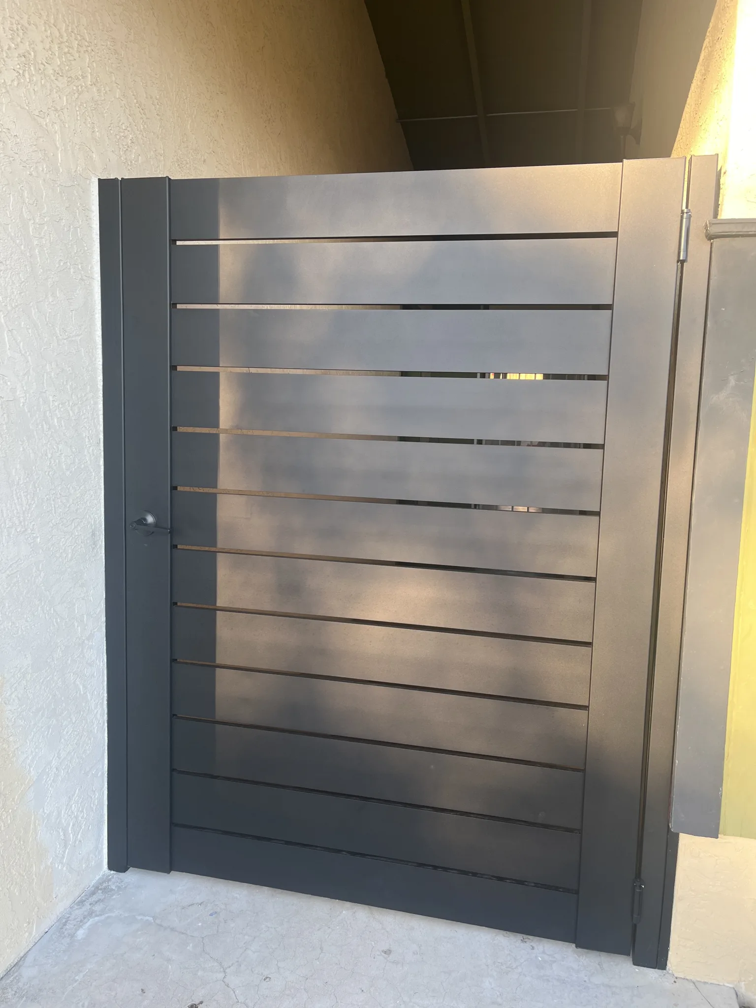 Dark gray pedestrian gate with horizontal slats next to a beige stucco wall
