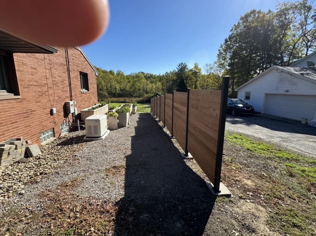 Long view of custom-style fence with Alumission aluminum posts and wood slats beside brick house and driveway.