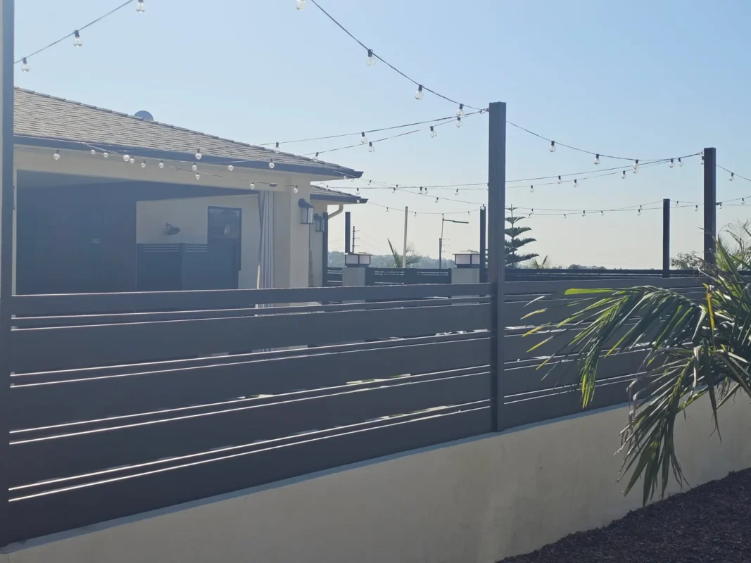 Close-up view of black horizontal slat aluminum fencing and posts with decorative lighting atop a low concrete wall