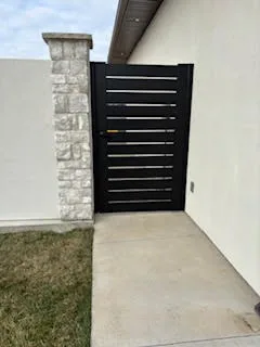 Black aluminum pedestrian side gate with horizontal slats between stone pillar and house wall, closed view