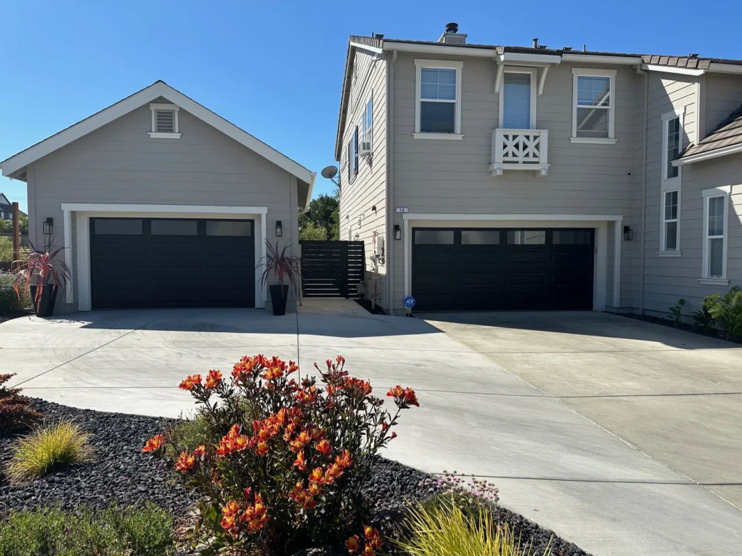 View of a residential driveway area with two black garage doors and a black horizontal slat side gate between two buildings.