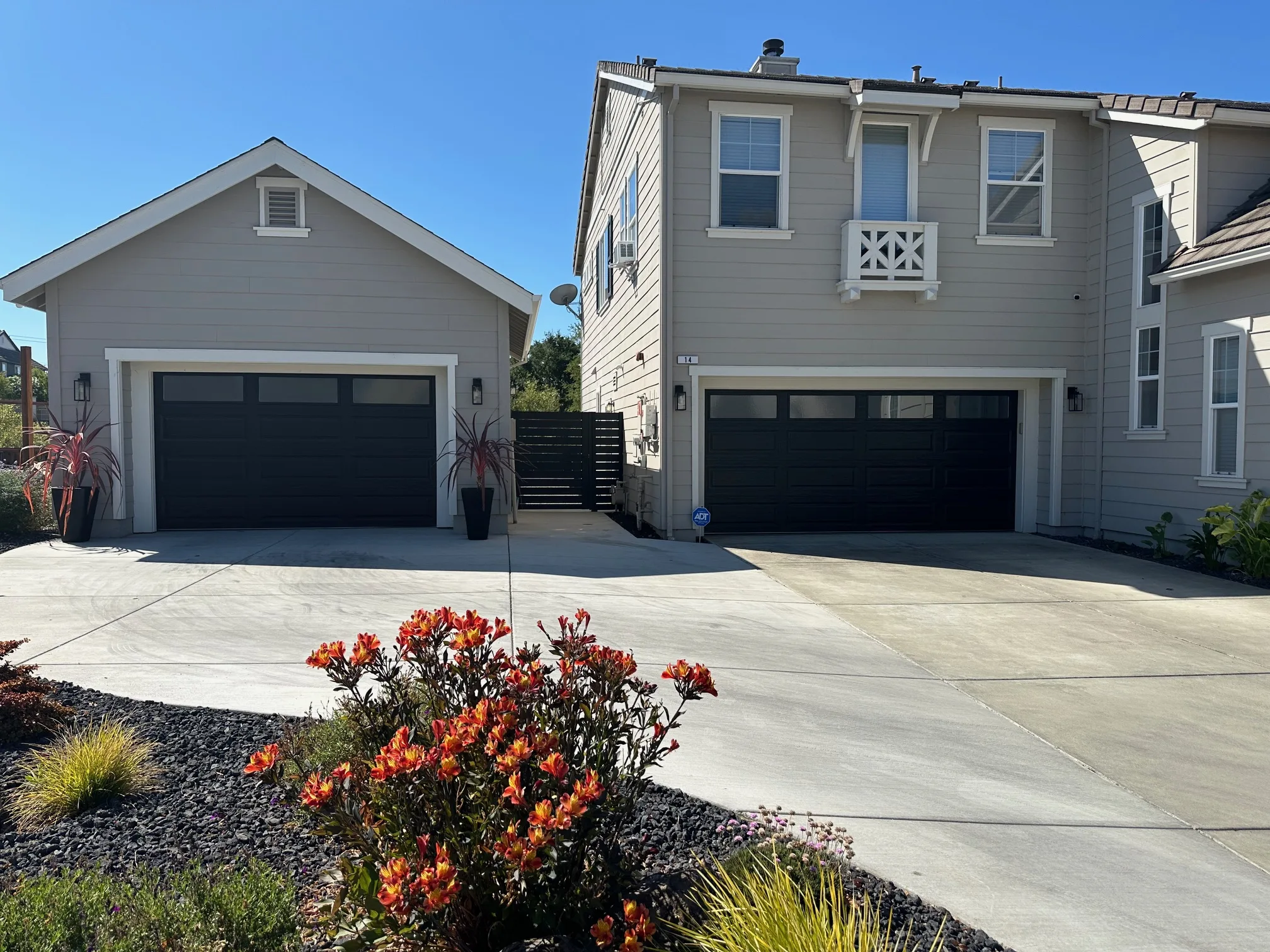 View of a residential driveway area with two black garage doors and a black horizontal slat side gate between two buildings.
