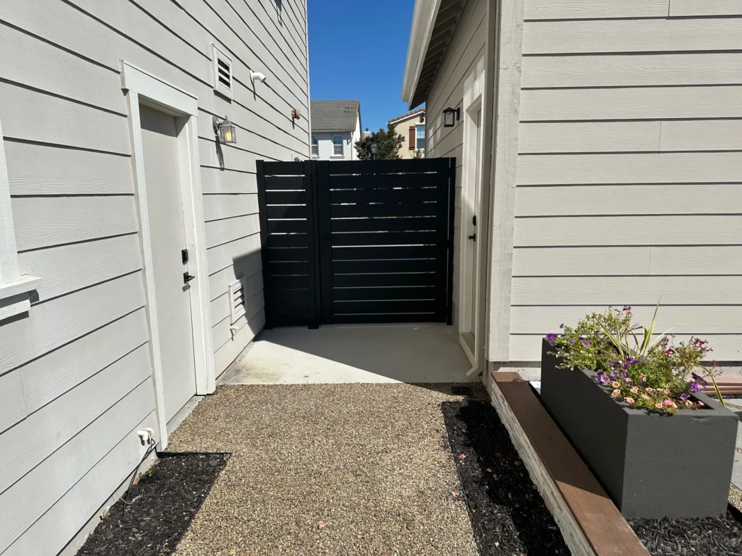 View of the black side gate enclosing a side pathway between residential buildings with utilities on the wall.