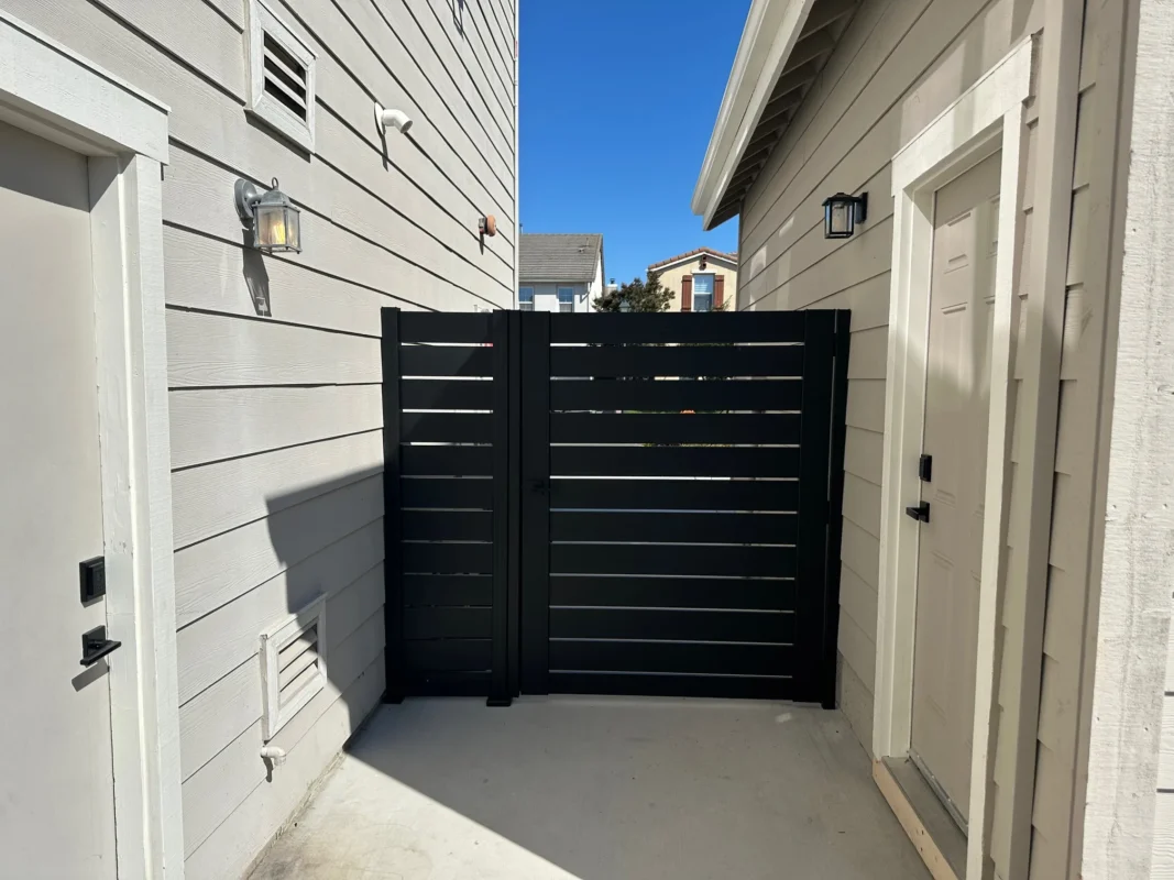 Close-up view of a black side gate with horizontal 5.5 inch wide slats and 1/2 inch gaps installed between two light-colored buildings.